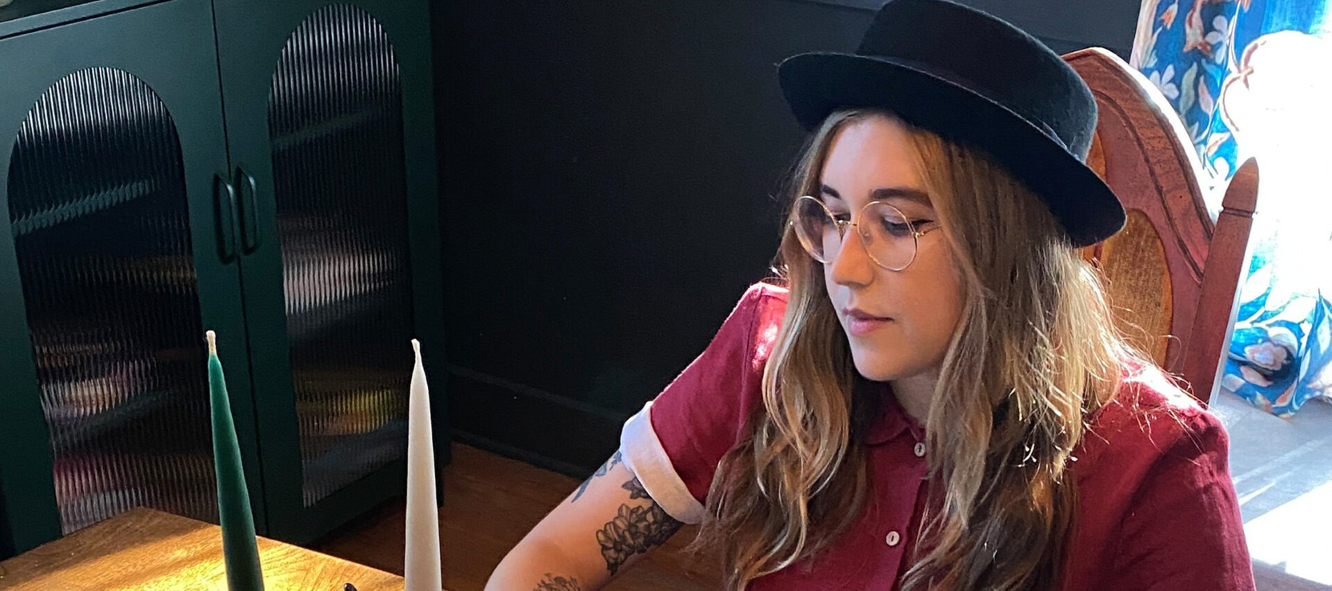 A young woman with light skin tone sits at a wood table and appears to be drawing on a small rectangular piece of paper. She wears a black hat and button-up shirt. The room's walls are painted dark.