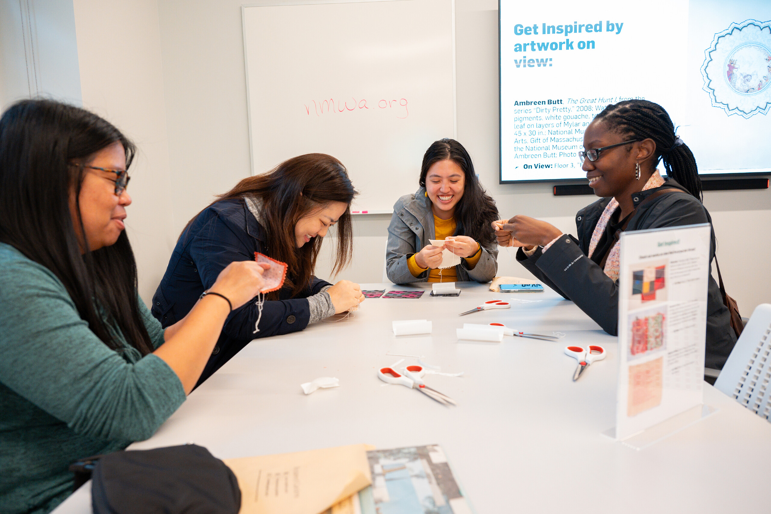 Four women sit at a table and hand-stitch white thread into white pieces of fabric.