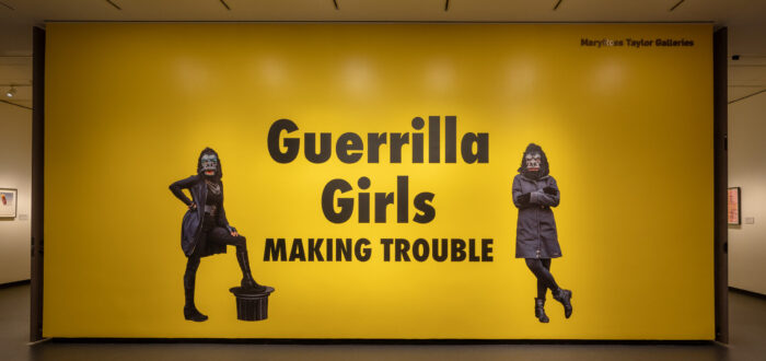 A large yellow wall with black text that says "Guerrilla Girls Making Trouble." On both sides of the text is an image of a figure wearing all black and a gorilla mask.