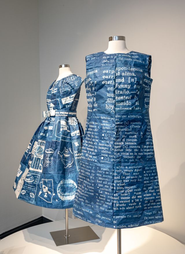 A sleeveless rich blue short dress is placed on a headless white dress form. The dress has white texts printed on the surface. Behind it is another, similar dress with short sleeves and belt.