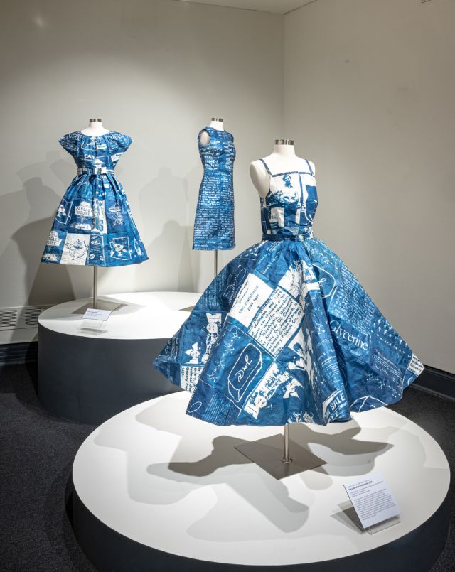 A group of three rich blue and white dresses placed on a headless white mannequin on rounded pedestals. The dresses are printed with imagery and text.