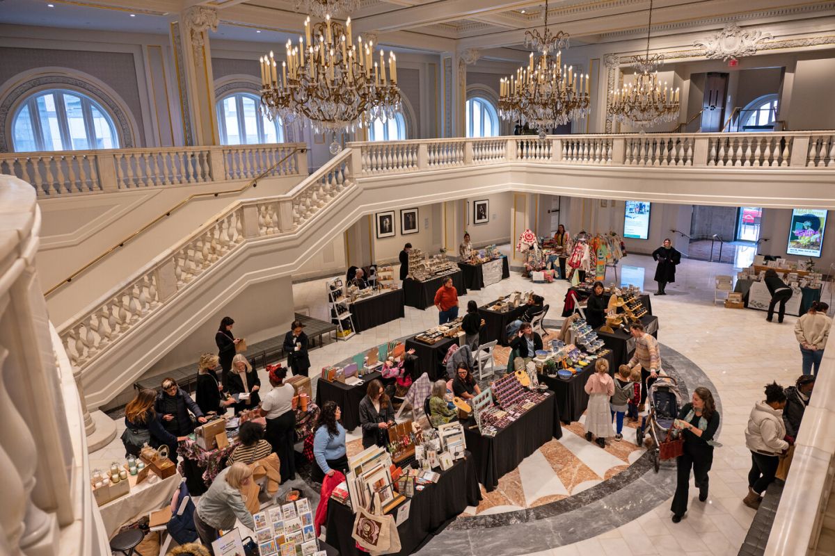 An elegant indoor market with vendors at tables displaying various crafts and goods. Shoppers are browsing under ornate chandeliers and a grand staircase, surrounded by white walls and large arched windows.