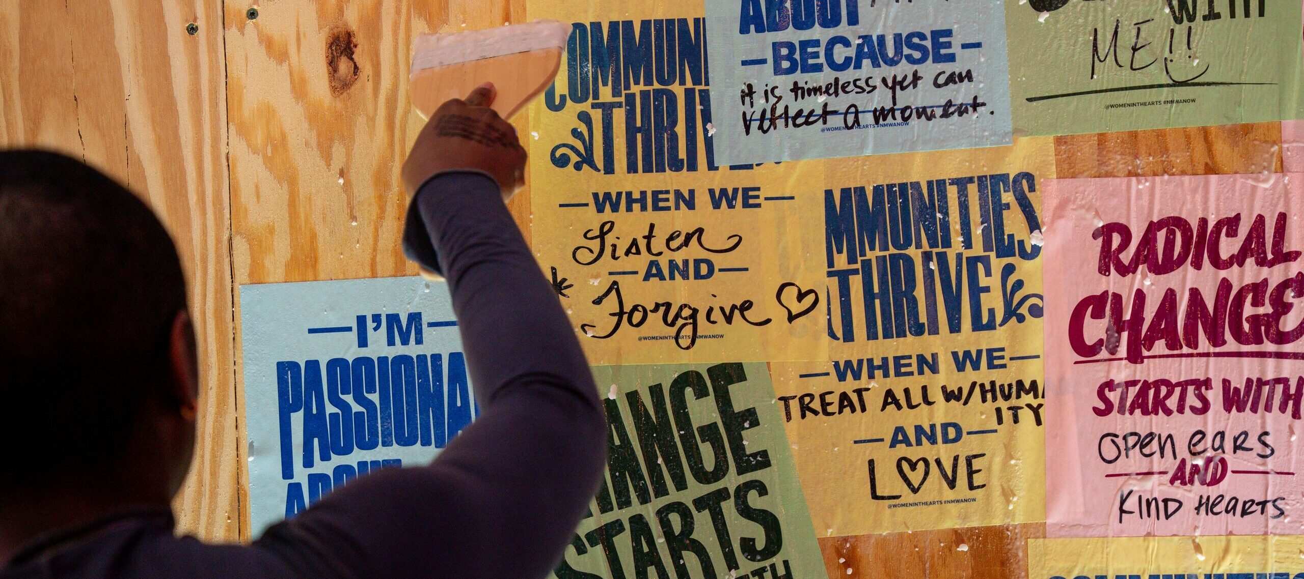 A woman uses a paint brush to paste colorful sheets of paper onto a wooden wall.