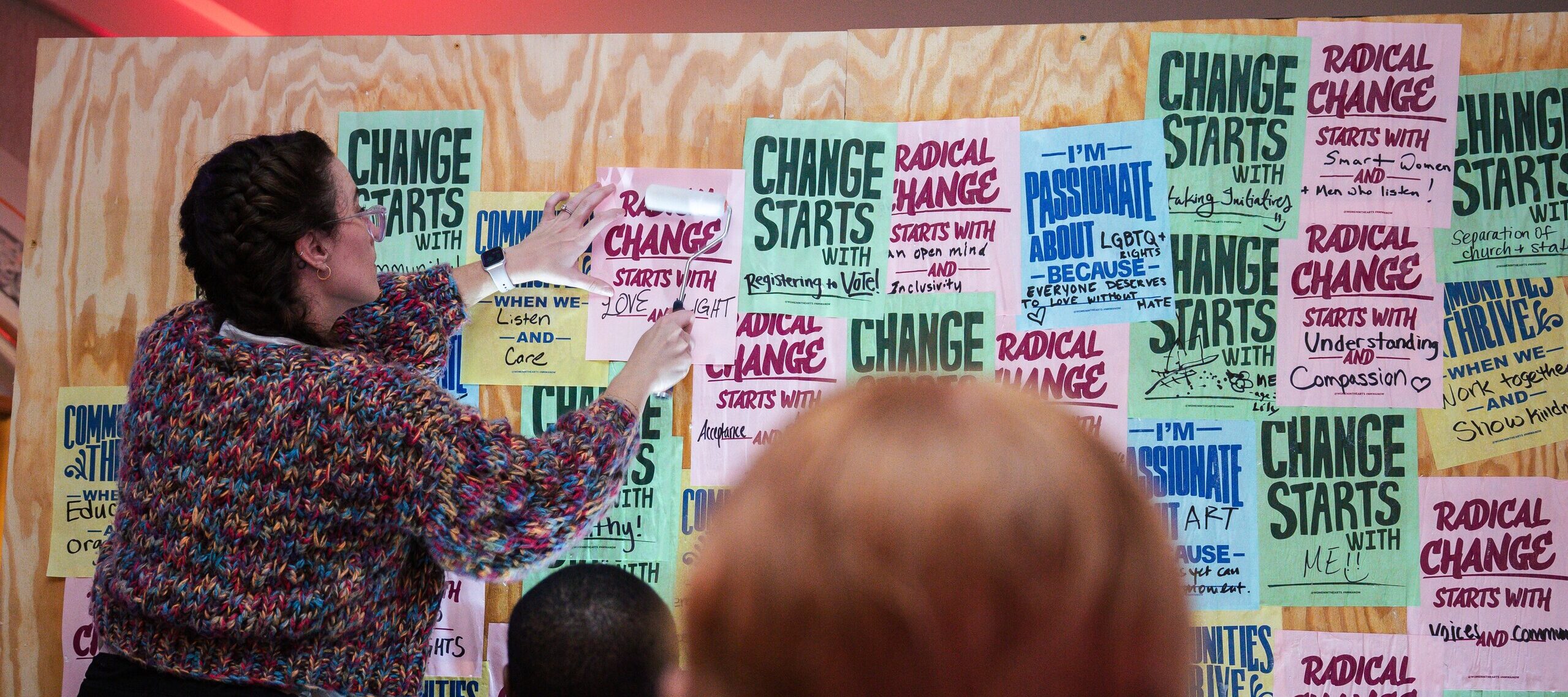 A woman pastes a paper with the text “Radical change starts with me!” onto a wall covered in colorful, handwritten posters with motivational messages, while two people look on from the foreground.