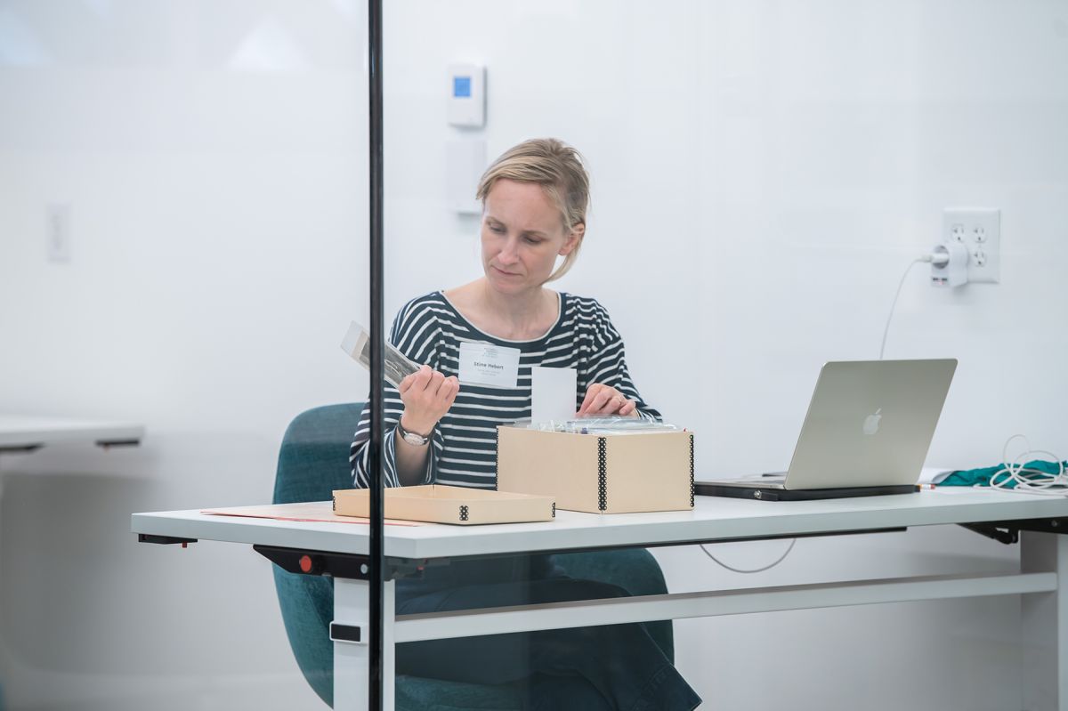 A woman with a light skin tone and blonde hair is sitting behind a glass wall by a desk. With one hand, she is holding a card and taking a closer look, with the other hand she is holding onto a box of cards.