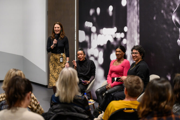 A woman stands speaking into a microphone while three people sit on a panel beside her. The seated individuals listen and engage with the audience. The background features a large black-and-white photo. Audience members are seated in the foreground.