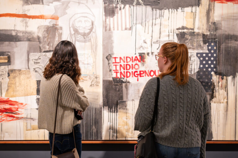 Two female visitors in grey sweaters look at an abstract painting. The painting is primarily beige and grey blocks of color, and the words "Indian" "Indio" and "Indigenous" are written in red paint.