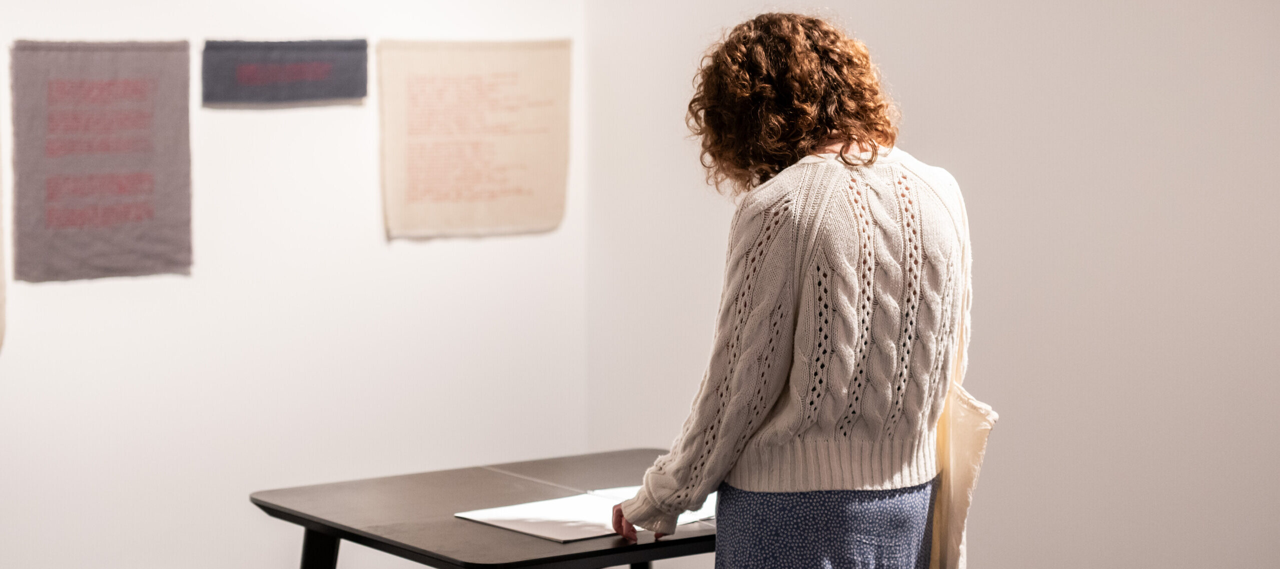 A person with curly brown hair, a white cable knit sweater, and a blue skirt reads a booklet on a black table, in front of an art work of grey and beige cloths installed on the wall.