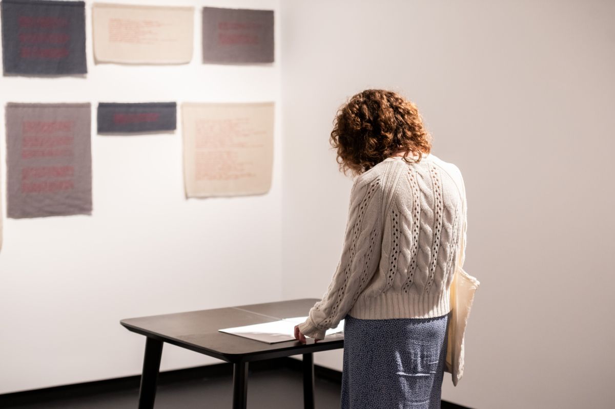 A person with curly brown hair, a white cable knit sweater, and a blue skirt reads a booklet on a black table, in front of an art work of grey and beige cloths installed on the wall.