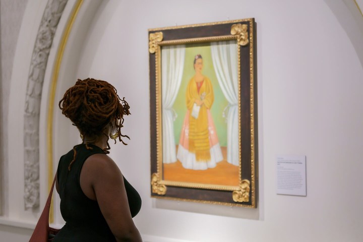 A person looks at a painting of a woman in a yellow, white, and pink dress.