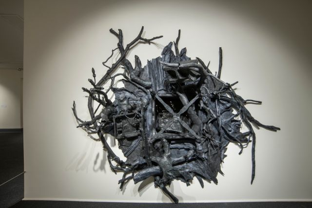 A contemporary black wall sculpture in a rounded shape with many twisted branches protruding out from the work. A void space is in the center. 
