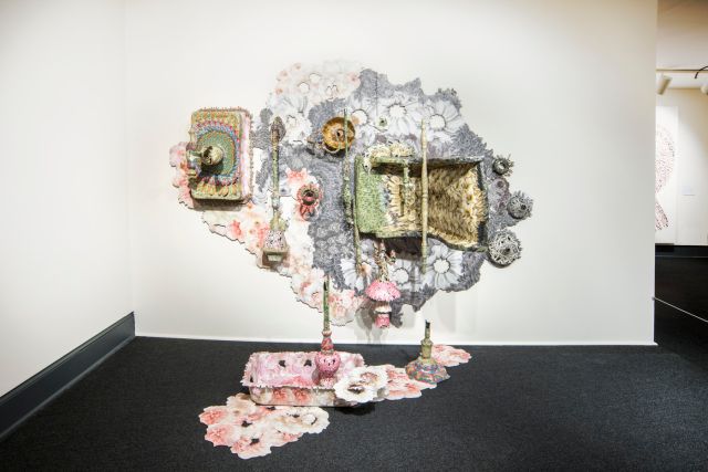 A contemporary wall sculpture with cut wallpaper piece pasted onto the wall and objects and home furniture protrude from the wall and onto the ground covered in colorful floral wallpaper. 