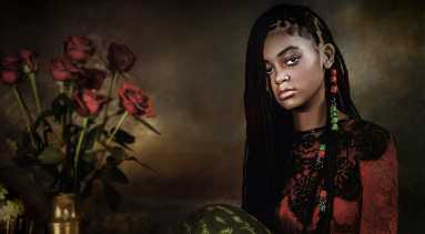 A seated girl with dark skin tone wears red and green and holds a watermelon. A vase of flowers is on a table to the left.
