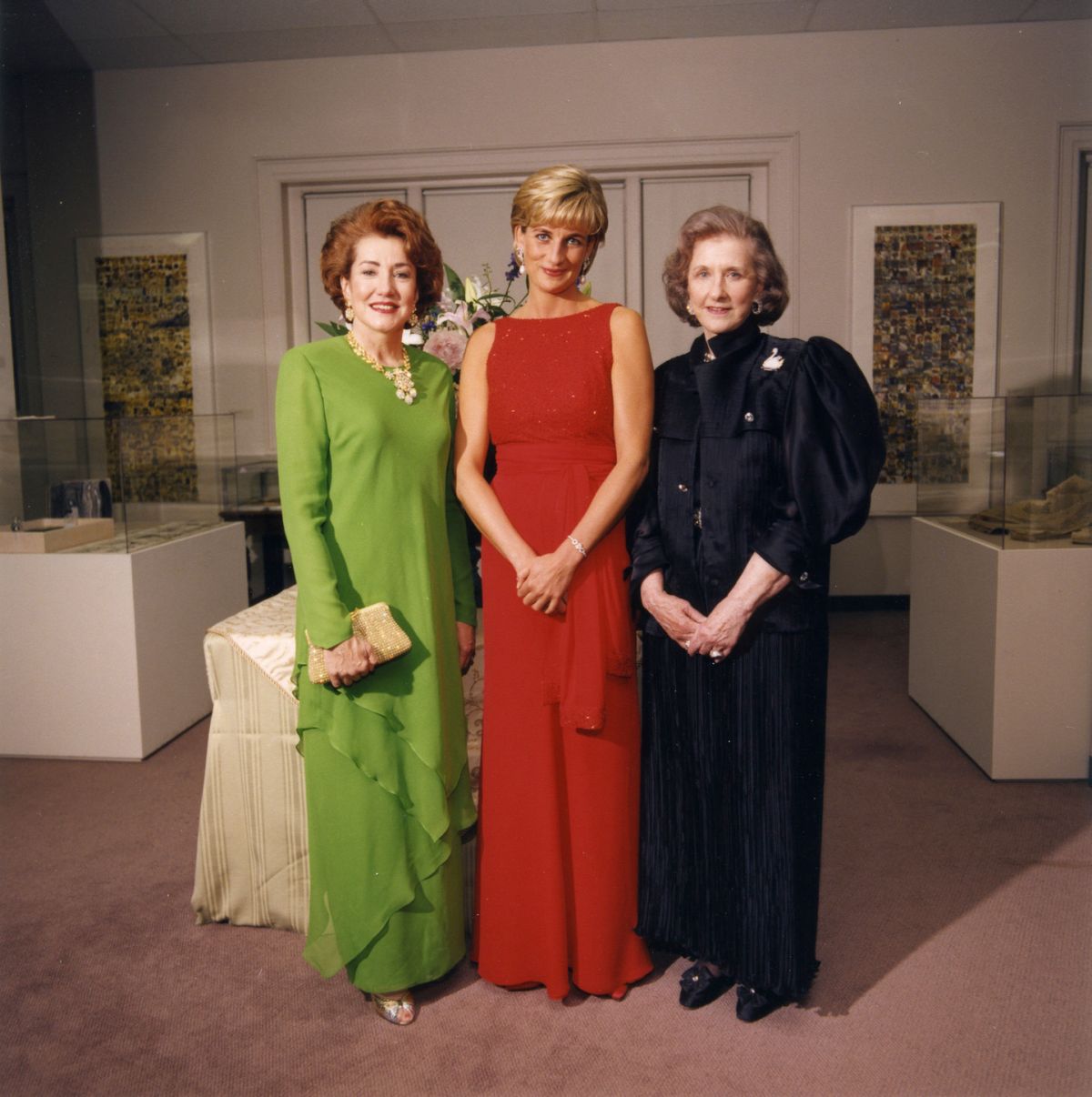 Three light-skinned women stand next to each other in fancy evening dresses inside an art gallery. The woman on the left wears a green dress, has red hair, and clasps a purse in her left hand; the women in the middle has short blonde hair, wears a red dress, and clasps her hands together in front of her; the women on the right has greying brown chin-length hair and wears a black dress, and also clasps her hands together in front of her. They all smile at the camera.