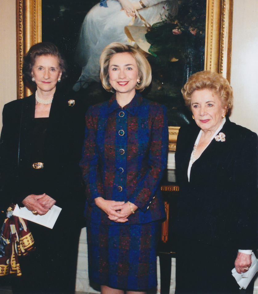 Three light-skinned women wearing formal skirt and blazer sets stand in a row smiling for a photograph. A woman with blonde hair wears a blue and red/orange striped suit, while the others are all in black with different broaches adorned on their left lapel