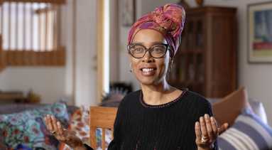 A dark-skinned women sits in a living room wearing a colorful headscarf knotted at the top; large, dark-framed glasses; and a long-sleeved black sweater. Her arms are stretched out at her sides with her palms facing up.