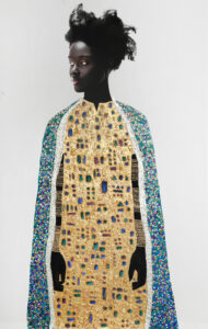 A photograph depicting a woman with dark skin and natural hair standing against a light background, wearing a cape and robe adorned with intricate gold patterns and colorful gemstones.