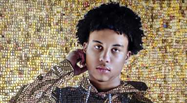 A mosaic style painting of a young brown skinned boy with black curly hair, who stands facing the viewer. He is wearing a brown hoodie sweatshirt, with the image of a parent holding their child on it. He and the background behind him is created from an elaborate layered mosaic of largely golden and brown hued square tiles.