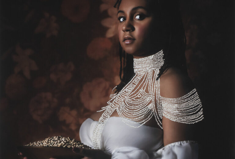A photograph of a Black woman wearing a long white gown. She is seated and her face is towards the viewer. She holds a plate of black-eyed peas.