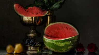 A still-life style photograph depicts a wedge of watermelon resting in a golden goblet. The remaining watermelon sits on the table to the right. Two lemons and blueberries are partially hidden near the base of the goblet.