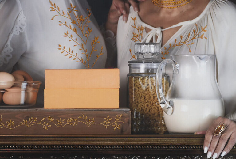 A photograph of two women in embroidered white tops stand behind a table with eggs, orange boxes, pasta in a jar, and a glass pitcher of milk, set against a dark background.
