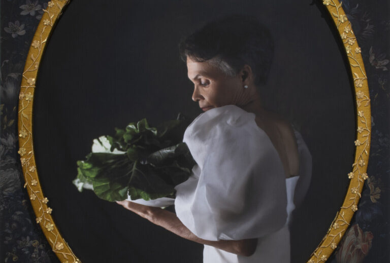 A photograph of an older Black woman with short grey hair in profile. She wears a long, white, gown with puffy sleeves. She holds and gazes at a head of collard greens.