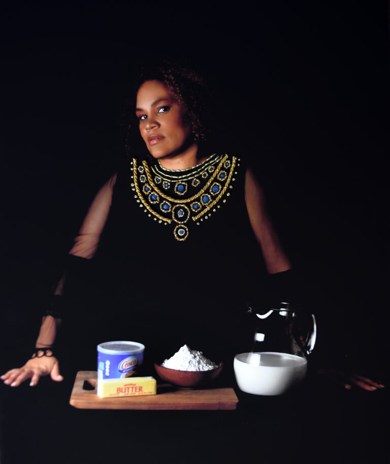 In a photograph, a woman with medium skin tone and dark, curly hair stands in front of a bowl of flour, butter, Crisco, and buttermilk. She wears a black dress, and gold thread and blue beads are stitched onto her clothes on the surface of the photograph.