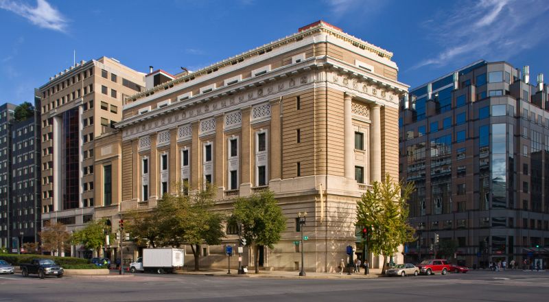 View of the museum from outside showing the Neoclassical building from one corner. The building is a tan-colored stone with an arched doorway, long vertical windows, and detailed molding around the roof.