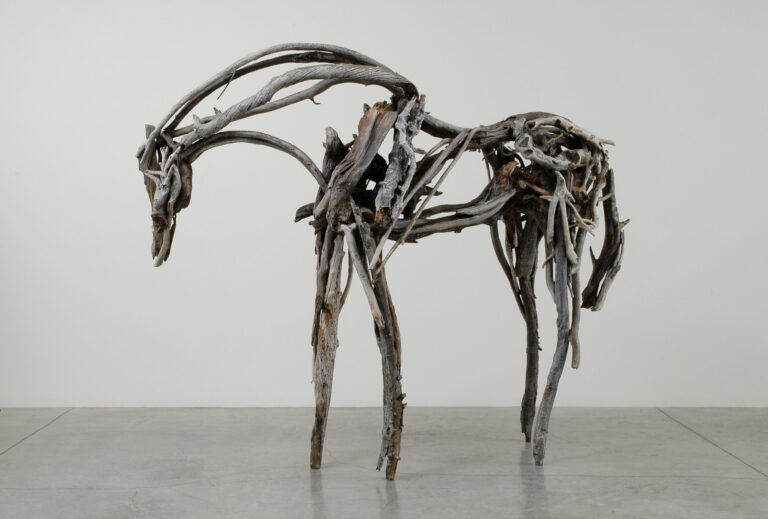 In this life-size sculpture, a horse stands facing left with its neck and head bent down. The horse appears to be made of many sticks of driftwood that are different shades of gray, different textures, and different lengths, all coming together to form a sculpture that evokes a horse’s movement.