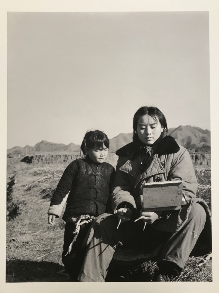 In this black-and-white photograph, a young woman of Asian descent sits next to a child of Asian descent in a rural, mountainous setting. They wear coats and look down at an object in the woman’s hand.