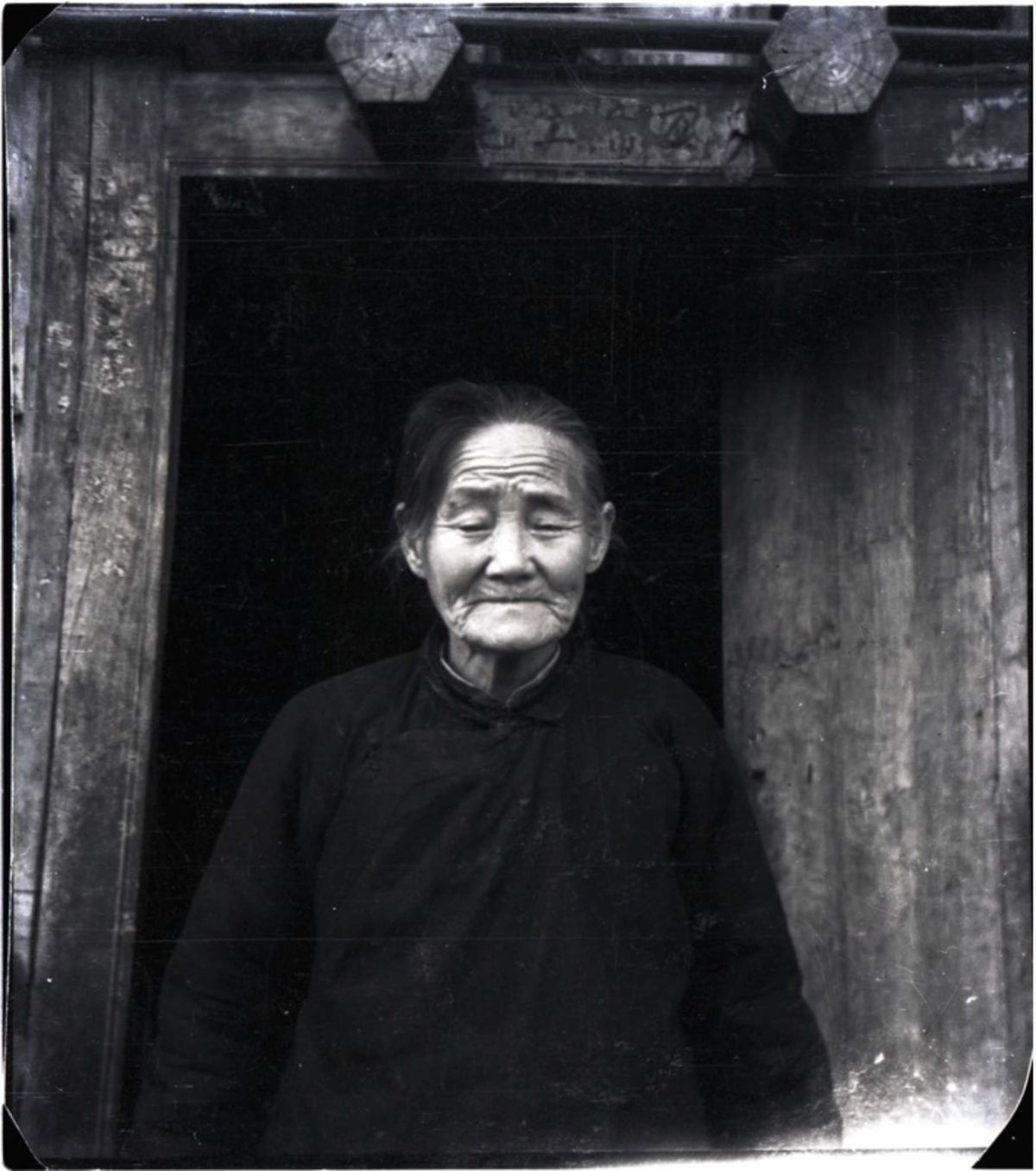 A black-and-white image shows an elderly Asian woman in dark attire standing in a wooden doorway.