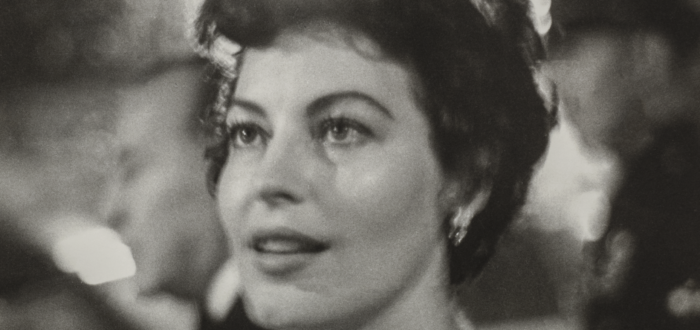 Ava Gardner, a woman with light skin tone and wavy, short brown hair, is shown from the shoulders up. She wears a jeweled necklace and is surrounded by lights and people at a party. She looks to the side of the camera at something out of frame with a fixed gaze and slight smile.