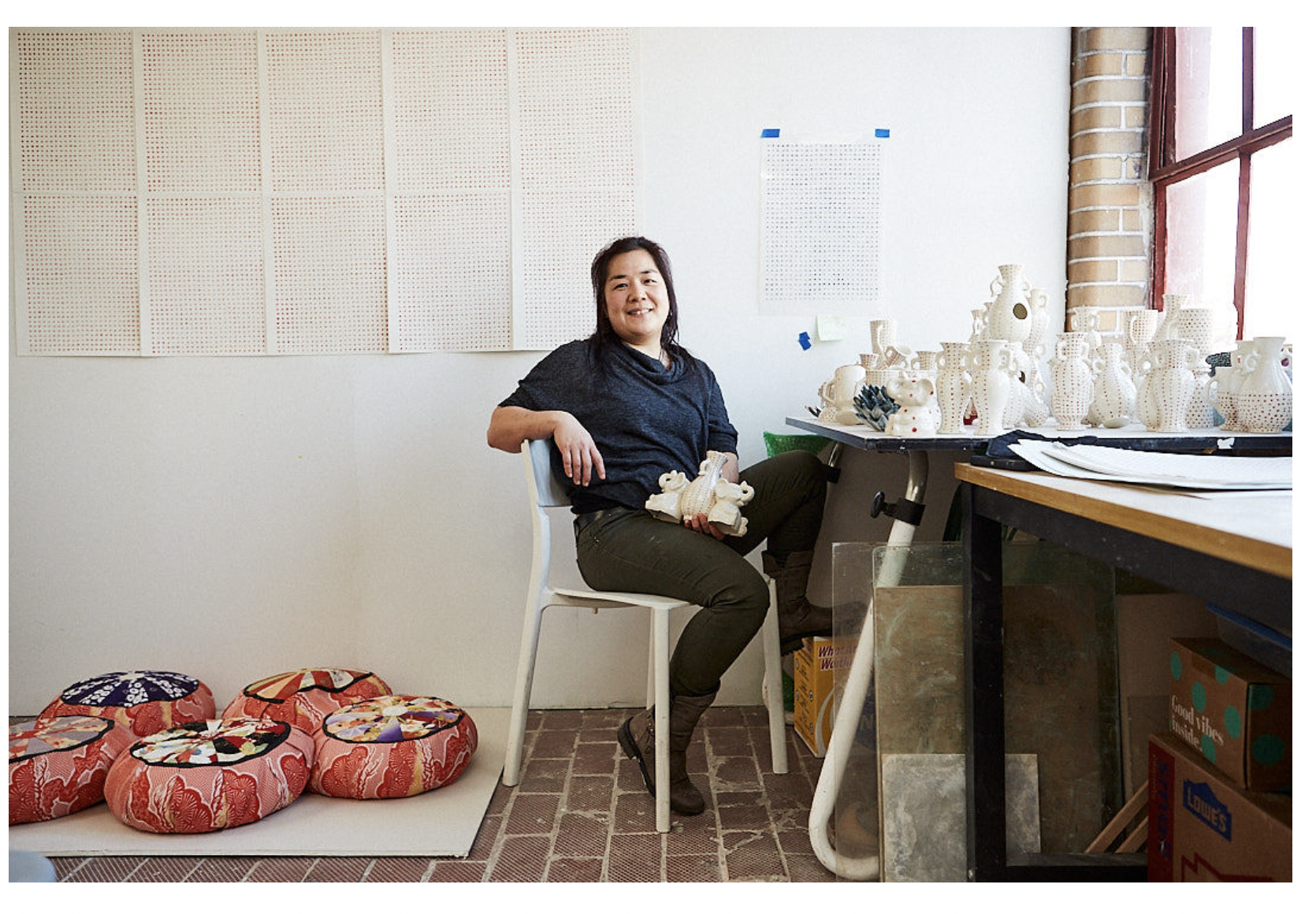 The artist sits in her studio, leaning back on a chair and holding one of her sculptures. She has long, black hair and a light skin tone, and she wears a blue sweater and olive green pants. The studio is very bright, with a white wall, several pink and red pillows scattered on the floor, and a whole table full of white vases.