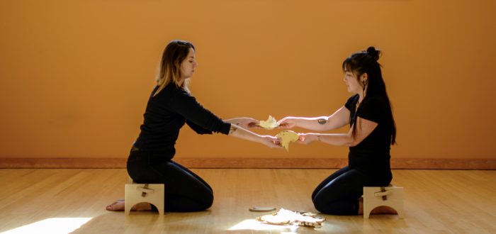 Two light skinned individuals sit on short wooden stools facing one another. They each hold a bird-shaped figure in one hand and are passing it to the other.