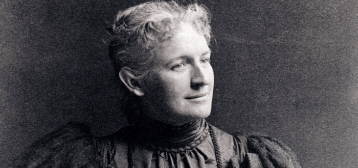 A black-and-white portrait photograph of a light-skinned woman from the late 1800s.She wears a dark, ruffled dress that has a high collar and puffy sleeves. Her light hair is pulled back and she looks to the right, slightly smiling, while one hand rests on her hip and the other in her lap, atop a small leather bag.
